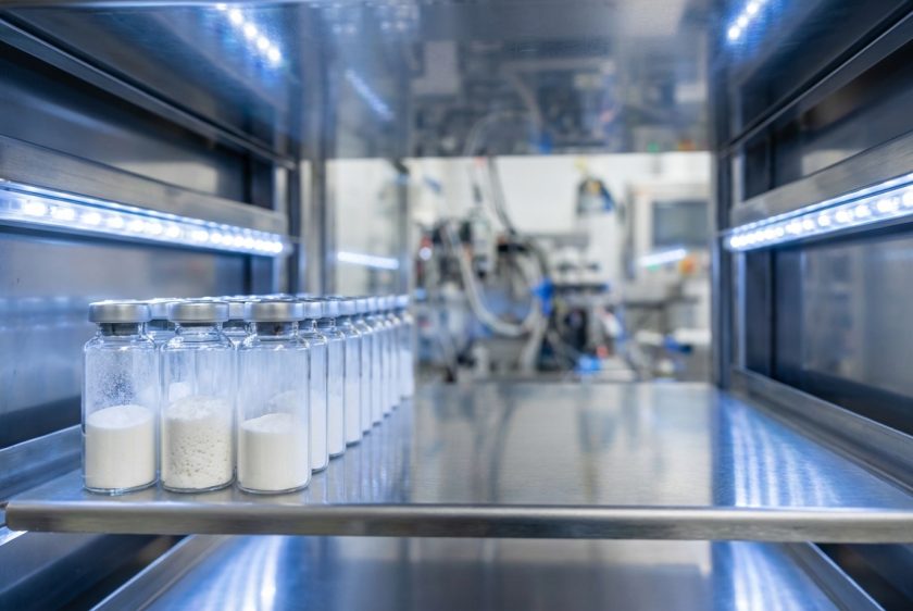 Modern bir liyofilizatör makinesinin içinde, raflara dizilmiş, beyaz toz halinde liyofilize edilmiş aşı veya ilaç içeren cam şişeler.