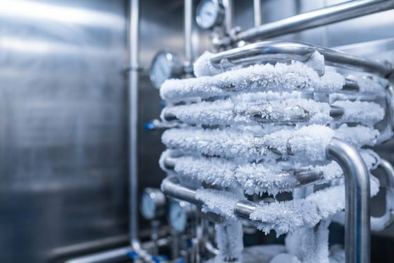 Bir liyofilizatörün kondenser ünitesindeki paslanmaz çelik serpantinlerin üzerinde yoğun bir şekilde birikmiş beyaz, kristalize buzun yakın çekim fotorealistik görüntüsü.